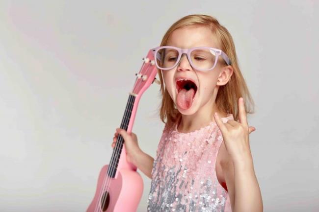 Rock and roll music concept. Cute caucasian little girl shows her to<em></em>ngue in a bright pink dress and glasses are playing the ukulele on a gray studio background with copy space