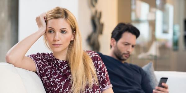 Young woman getting bored while man using phone in the background. Beautiful young woman feeling annoyed as man texting on phone. Young woman after an argument with her boyfriend in their living room.