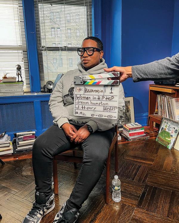 photo of seated short-haired woman with large glasses by bookcases in room painted electric blue with arm reaching in front holding film clapboard for Netflix docu<em></em>mentary