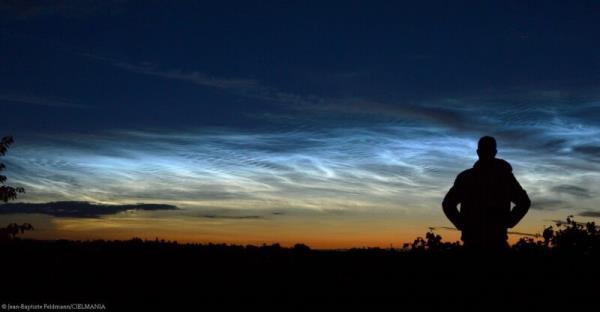 Photo of the horizon and sky. There is a man with his hands inside his pockets looking at the horizon, which looks orange. Blue sky. Whitish clouds above the horizon.