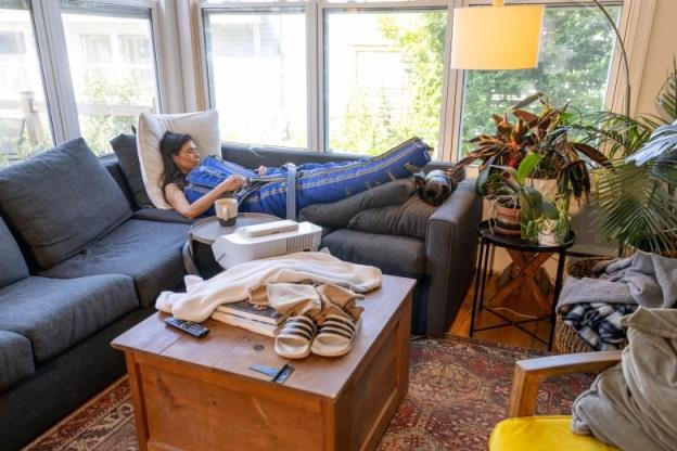 A woman is lying on a grey sectio<em></em>nal couch. She is in a blue suit that looks like a blanket. 