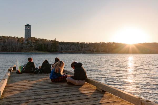 Young kids on a dock over water, tower in the distance and sun setting over treeline.