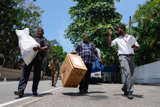 Three men, one carrying a cardboard box and another carrying a white package, walk together in a row down the street on a sunny day.