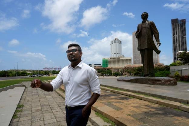 A man in a white button-down shirt and black glasses stands in a public square on a sunny day.