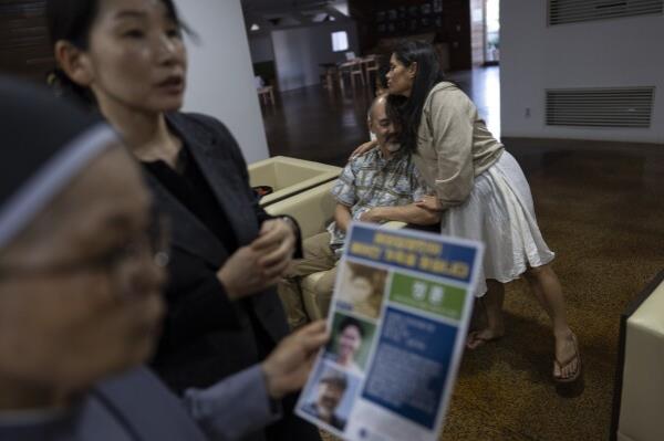 Kenneth Barthel, who was adopted by a single parent in Hawaii at 6 years old, is hugged by his wife, Napela, at the Sisters of Mary in Busan, South Korea, Friday, May 17, 2024. In the foreground, Sister Bulkeia, left, and Paek Kyeong-mi from Global Overseas Adoptees' l<em></em>ink discuss a flyer designed to uncover the details of Barthel's early life and find his birth family. (AP Photo/Jae C. Hong)