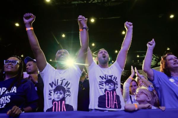 Two people in matching shirts lift their arms and cheer at a Kamala Harris rally.