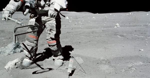 The hands, lower torso and legs of a spacesuited astronaut, on the gray moon, collecting rocks in a wire basket.