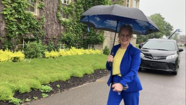 Woman in blue suit and yellow shirt, walking with umbrella and smiling at camera