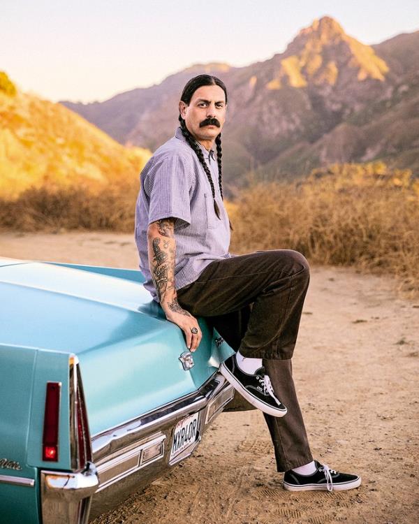 photo of man with black mustache and long black hair in two braids sitting on trunk of blue Cadillac with one foot on bumper, with mountains in background