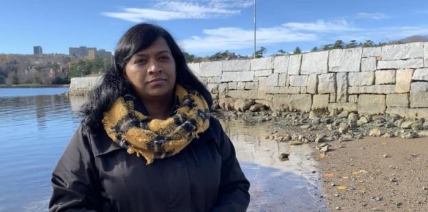 Woman looks at camera. Background is a calm blue ocean and a stone wall. 