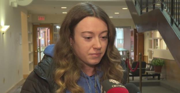 A young woman with long brown hair, wearing a dark hooded jacket, looks sad as she talks into two news microphones. 