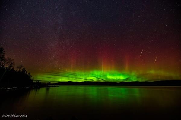 Green, yellow, orange and pink lights over the horizon. The lights are reflected in the water. There are 2 streaks on the right.