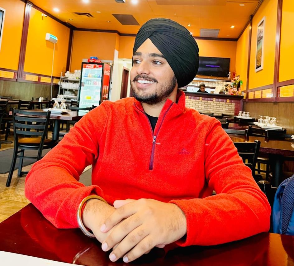 A man in a black turban and a red sweater sits inside a brightly lit restaurant, smiling and looking away from the camera.