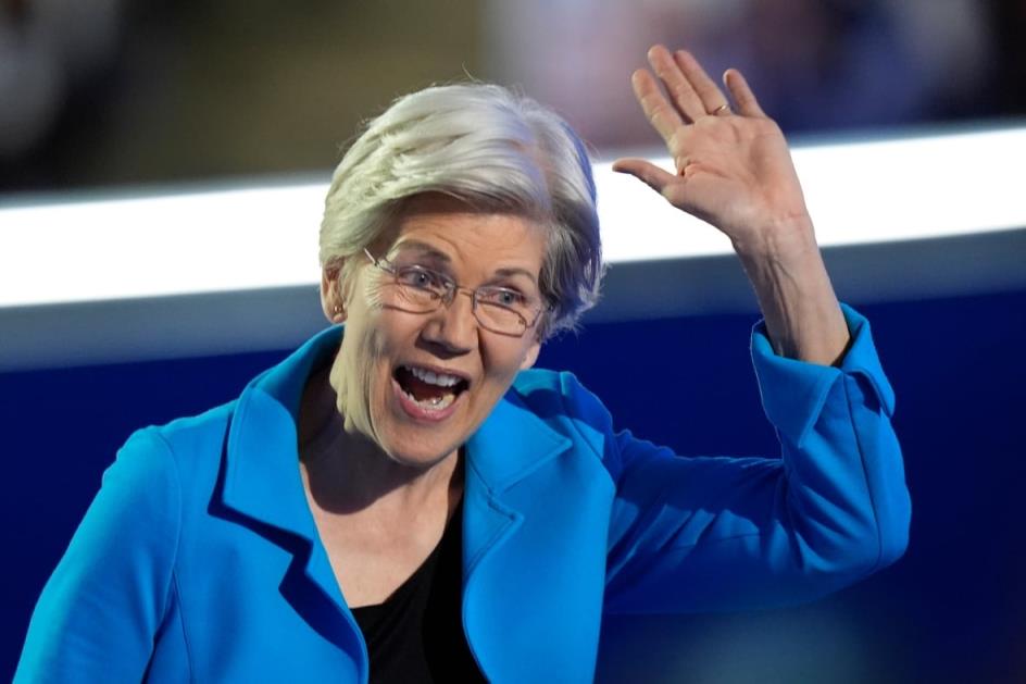 A woman with short, grey hair wearing a blue blazer smiles and raises her hand. 