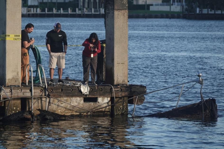 People standing on the edge of a pier survey the charred remains of a boat in the water.