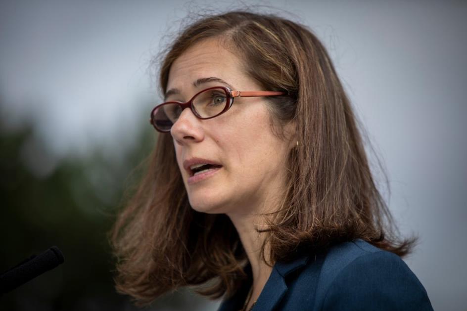 A white woman with brown hair and glasses is pictured speaking in profile.