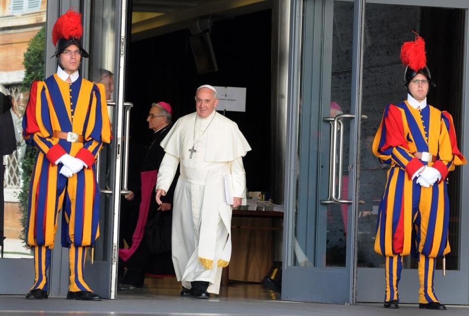 An elderly man in white walks through door with men in colourful dress and armour standing on either side of him.