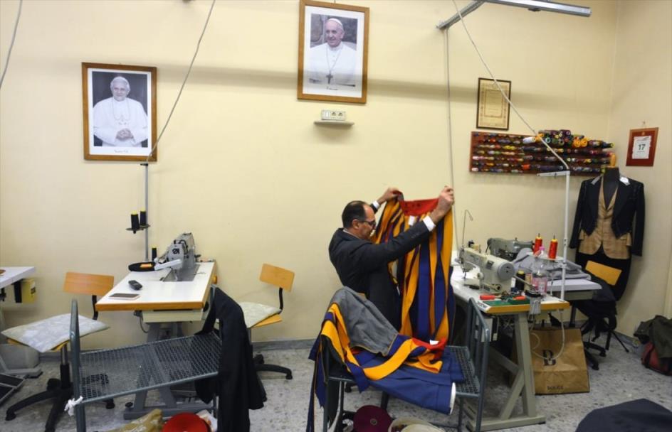 A man seated at a sewing machine holds up a yellow and blue striped garment.