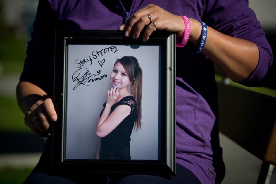 A woman holds a f<em></em>ramed photo of a teenaged girl with long brown hair wearing a black dress. The words 'stay strong' are written on the glass in Sharpie.