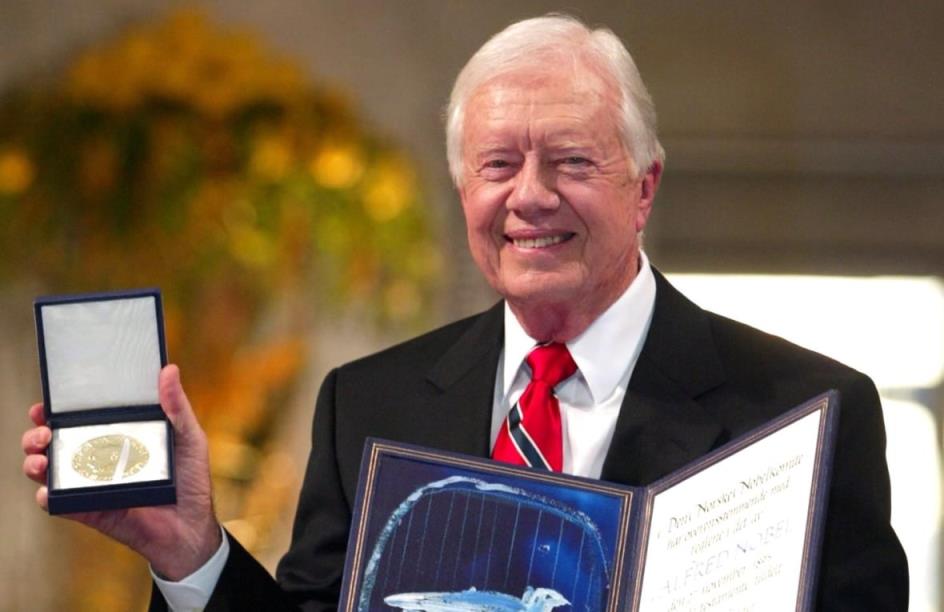 Wearing a dark suit, Jimmy Carter smiles while holding his Nobel Peace Prize.