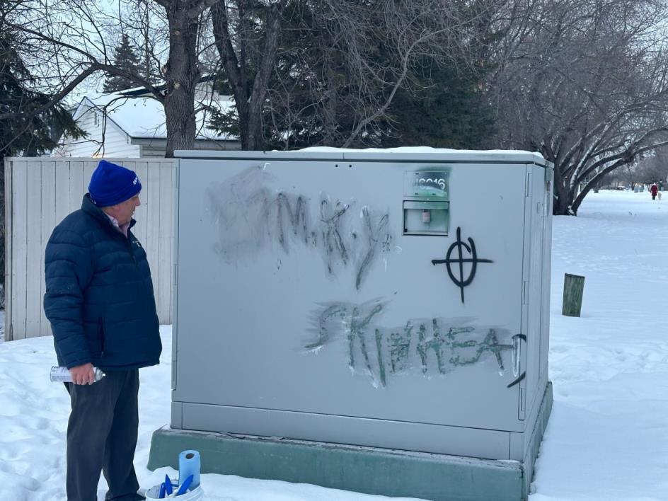 A man stands in front of some graffiti outdoors that's been partially deleted, with a swastika, the words 