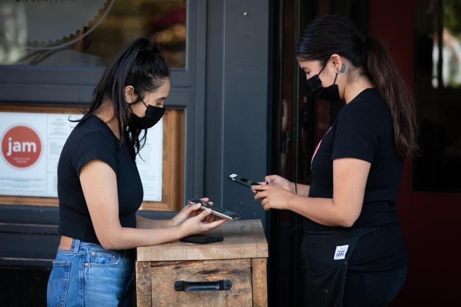 A masked woman scans another masked woman's phone at a restaurant entrance.