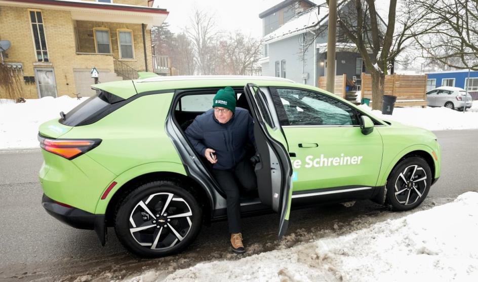 Mike Schreiner gets out of the rear of a green-painted electric vehicle, on a snowy street. 