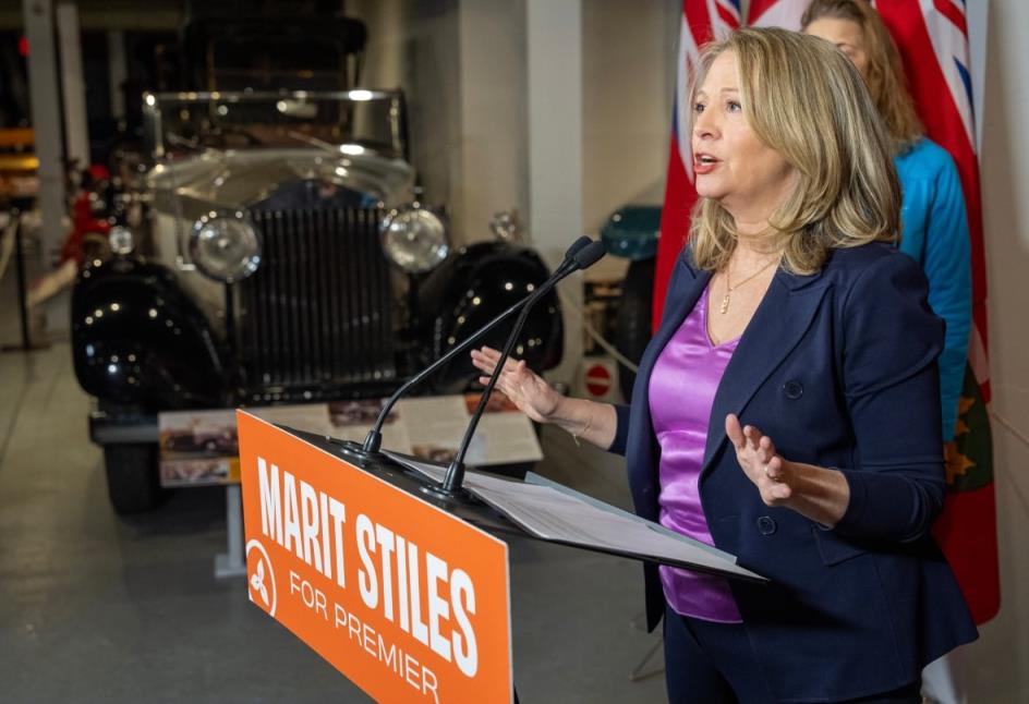 Marit Stiles, seen from the side at a podium, with an antique car in the background. 