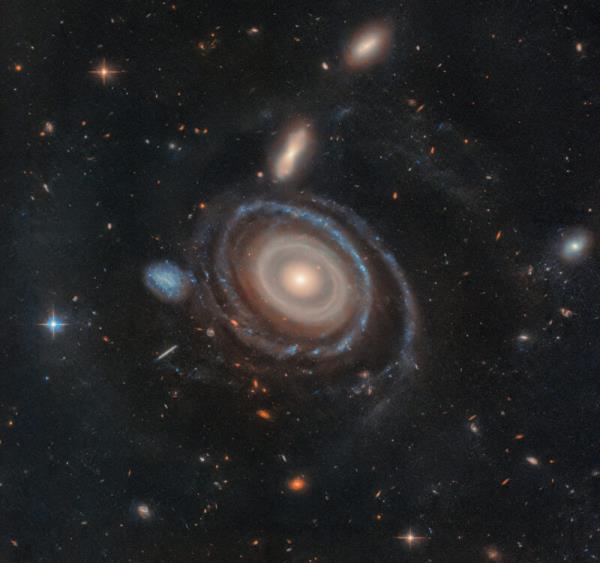 A galaxy with 9 rings in cream and blue to<em></em>nes and smaller galaxies in the background.