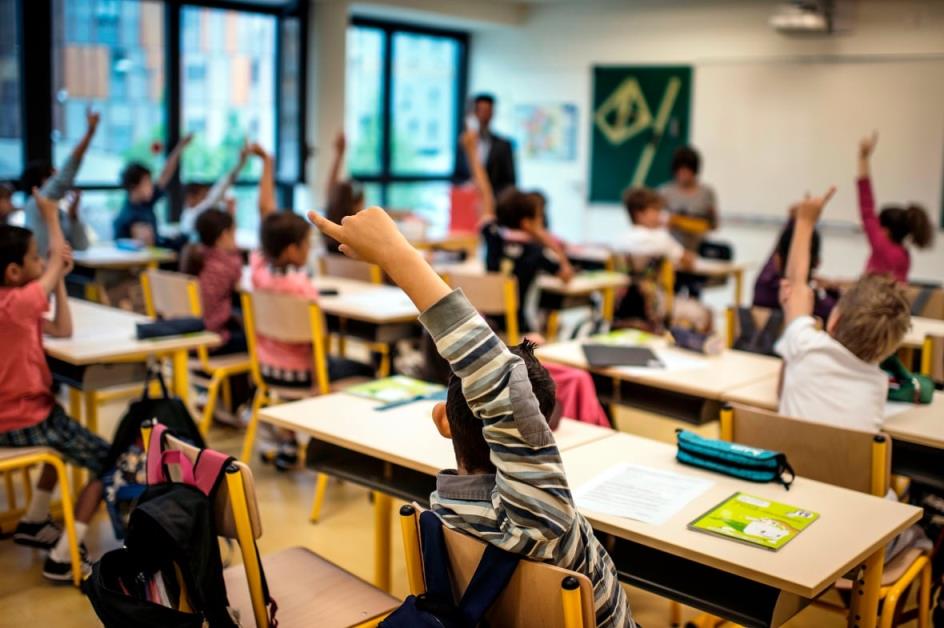 A classroom of elementary-aged students raise their hands in a classroom, as a male teachers stands by a set of windows in the background.