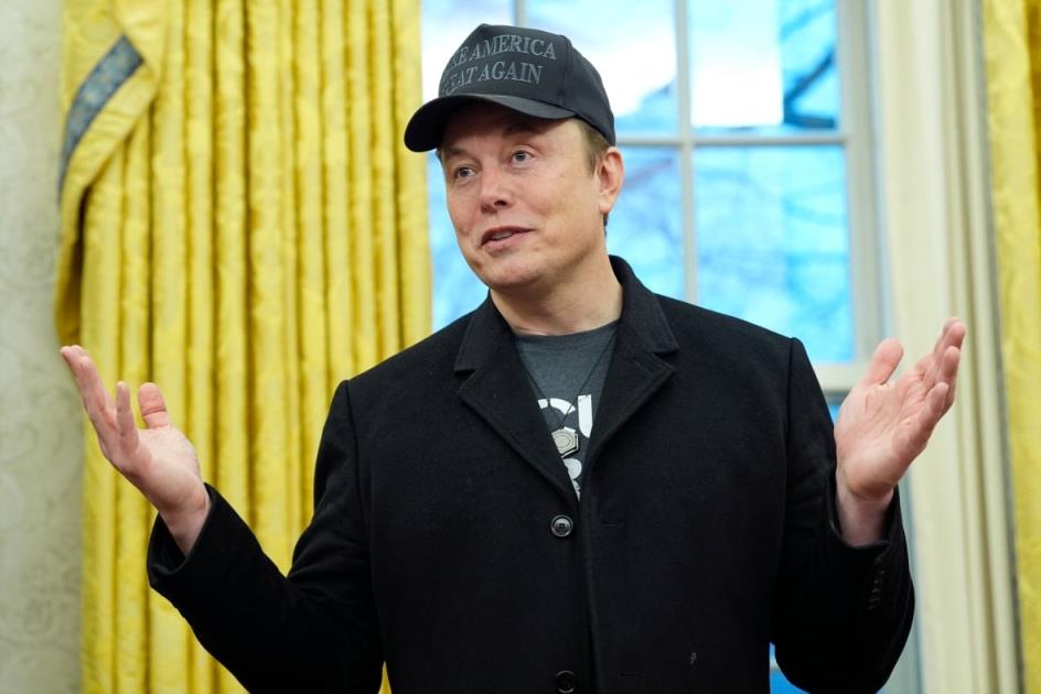 A cleanshaven man wearing a dark blazer, tshirt and ba<em></em>seball cap extends his arms while speaking indoors.