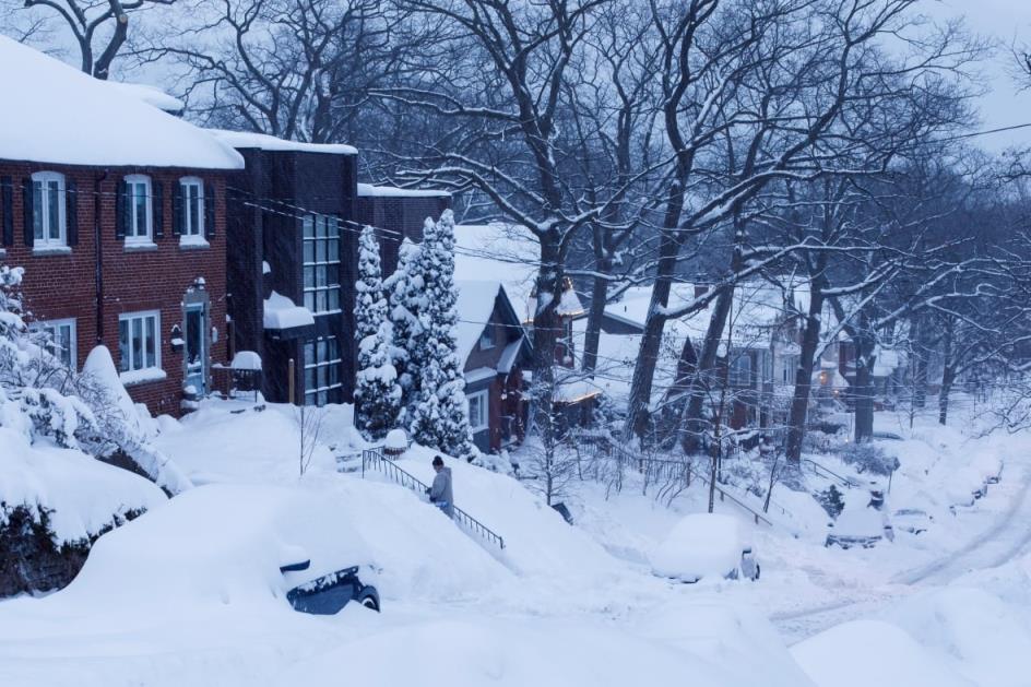 A person shovels front steps in east-end Toronto, while parked cars on the hilly street are buried by snow.