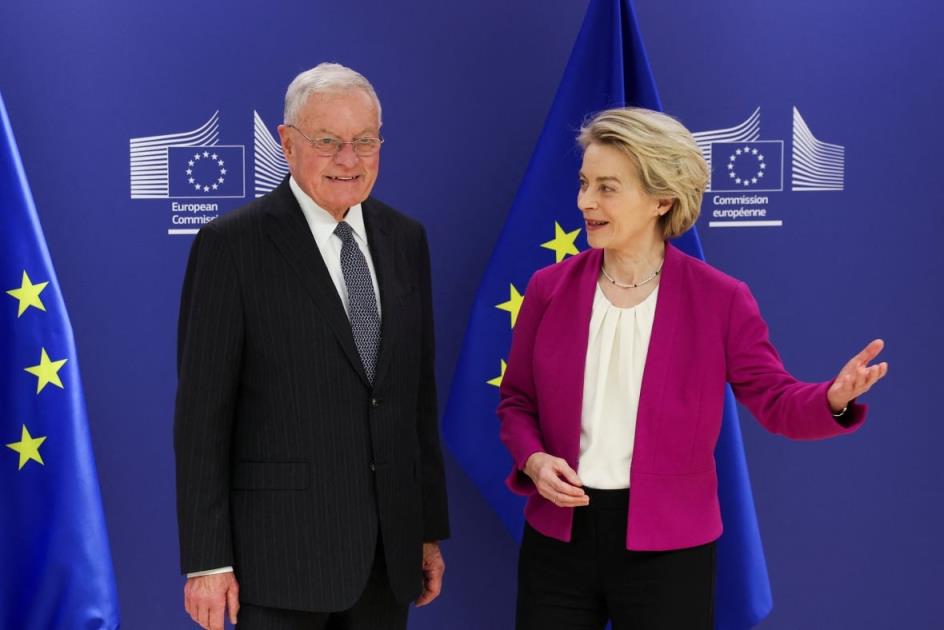 An older woman wearing a blazer and pants gestures with her hands while standing beside a white-haired, cleanshaven and bespectacled man wearing a suit and tie.