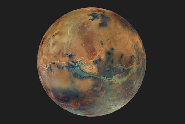 Mars is red: Reddish planet with darker regions on its rocky surface. A long canyon cuts across just south of the equator.