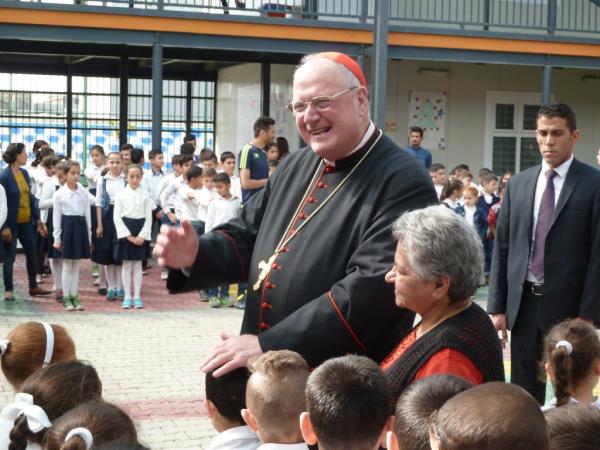 Cardinal Timothy M. Dolan in a red hat and black robe, greeting a teacher and students in the courtyard of the Al Bishara School