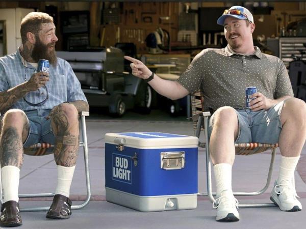 post malone, left, and shane gillis, in a super bowl commercial for bud light, both are seated in lawn chairs in front of a garage with a cooler between them